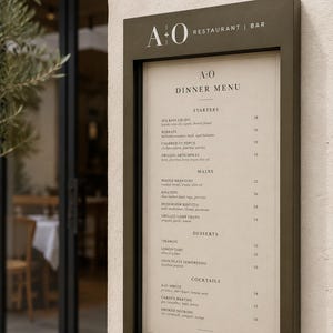 May include: A dark olive green rectangular menu display case mounted on a light beige wall. The sign features the text A+O RESTAURANT | BAR at the top and displays a printed dinner menu with sections for starters, mains, desserts, and cocktails.