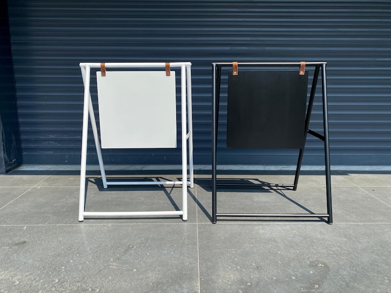 May include: Two A-frame sidewalk signs, one white and one black, with blank surfaces for displaying messages. Both signs have brown leather straps.