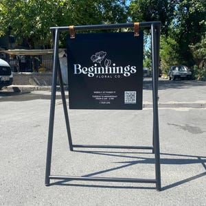 May include: A black A-frame sign with a black sign that says "Beginnings Floral Co." in white text. The sign also includes a QR code and information about weekly hours.