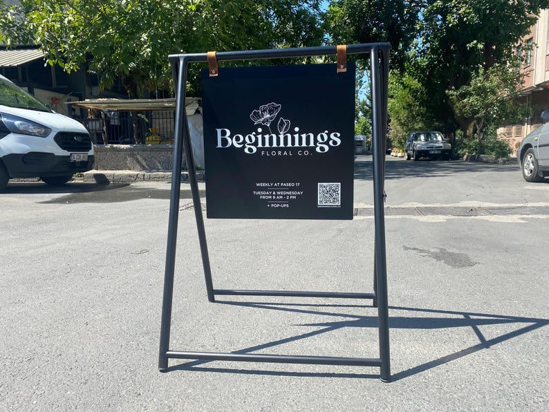 May include: A black A-frame sign with a black sign that says "Beginnings Floral Co." in white text. The sign also includes a QR code and information about weekly hours.