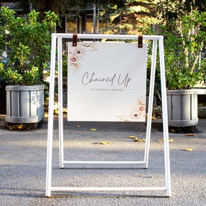 May include: White A-frame sign with a white background and brown leather straps. The sign says "Chained Up" in cursive script with the words "Handmade Jewelry" underneath. The sign has a floral design with pink and white flowers.