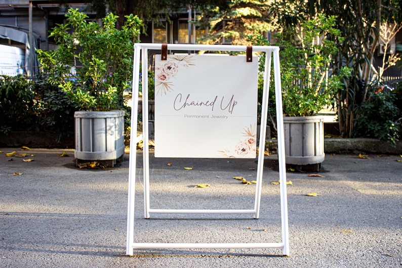 May include: White A-frame sign with a white background and brown leather straps. The sign says "Chained Up" in cursive script with the words "Handmade Jewelry" underneath. The sign has a floral design with pink and white flowers.