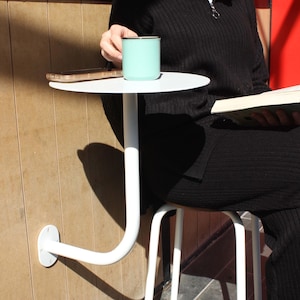 Op de afbeelding: Een witte, aan de muur gemonteerde tafel met een rond blad en een wit metalen frame. Een persoon zit op een witte kruk en gebruikt de tafel. Een turquoise mok en een mobiele telefoon staan op de tafel.