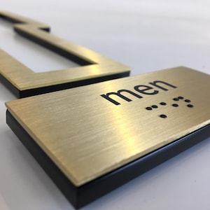 May include: Gold-colored metal sign with black lettering that reads "men" and braille dots. The sign has a black border.