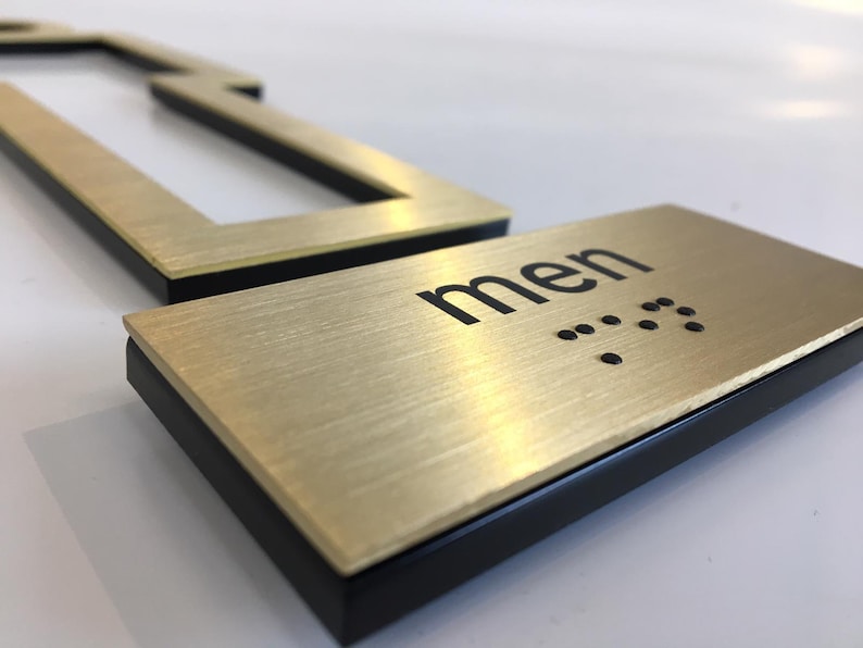 May include: Gold-colored metal sign with black lettering that reads "men" and braille dots. The sign has a black border.