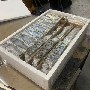 May include: A white styrofoam box with a lid open, revealing a set of gold-colored metal letters and numbers wrapped in plastic. The letters and numbers are arranged in rows and columns, and they are all the same size.