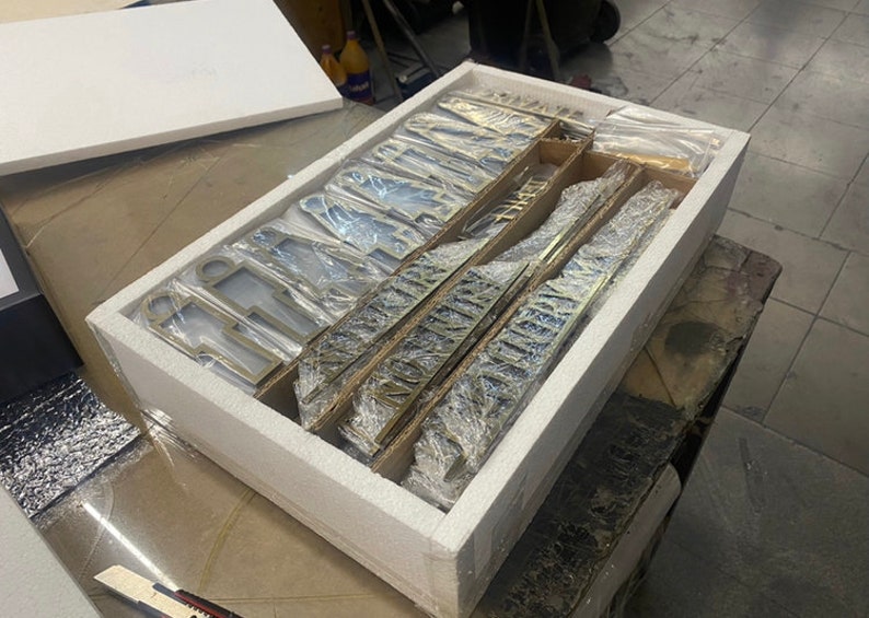 May include: A white styrofoam box with a lid open, revealing a set of gold-colored metal letters and numbers wrapped in plastic. The letters and numbers are arranged in rows and columns, and they are all the same size.