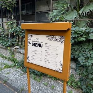 Op de afbeelding: Een bruin metalen bord met een witte achtergrond en zwarte tekst. Het bord zegt "Restaurant Menu" en somt verschillende eetwaren op met prijzen. Het bord is gemonteerd op een bruin metalen standaard.