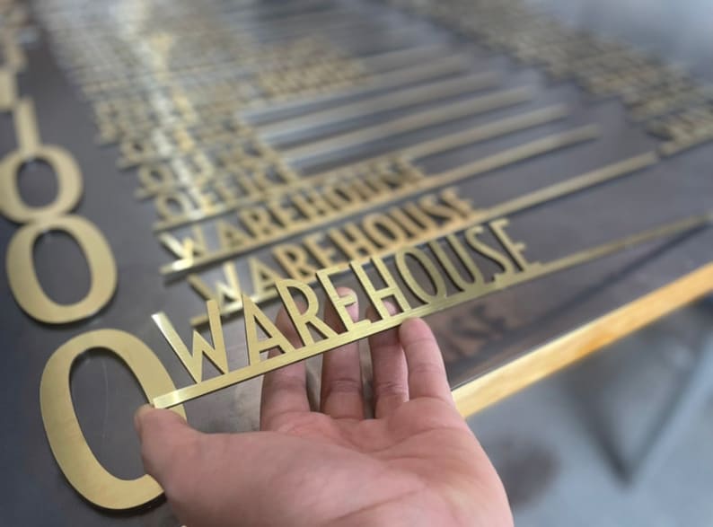 May include: A close-up of a hand holding a gold metal sign that reads "WAREHOUSE". The sign is one of many similar signs, all of which are arranged in a row.