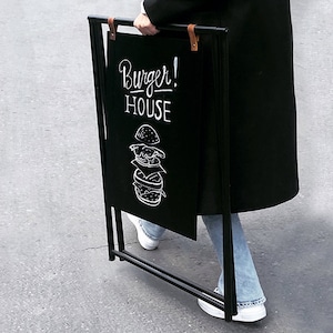 以下が含まれることがあります： 黒いチョークボードの看板に、白いハンバーガーの絵と「Burger! House」の文字が書かれている。看板は黒い金属製のスタンドに取り付けられている。
