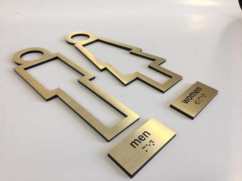 May include: Two gold metal signs with a stylized outline of a person. The signs are labeled "men" and "women" with braille lettering.