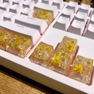Puede incluir: Teclado mecánico blanco y rosa con teclas de resina personalizadas. Las teclas son transparentes con flores amarillas y escamas doradas incrustadas. El teclado tiene un diseño estándar con teclas blancas y detalles rosas.