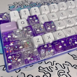 May include: A custom mechanical keyboard with white keycaps and a purple and clear resin spacebar and keycaps. The resin keycaps feature a celestial design with clouds, moons, and stars. The keyboard has a purple WIN key.