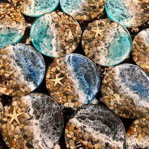 Könnte beinhalten: Ein Set runder Untersetzer aus Harz mit Strandmotiv. Jeder Untersetzer zeigt ein blaues und weißes Wellendesign mit weißem Schaum, braunen Kieselsteinen und einer weißen Muschel. Einige Untersetzer haben auch einen weißen Seestern.