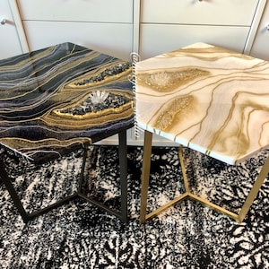 May include: Two hexagonal side tables with gold and white resin tops and gold metal legs. The table on the left has a black and gold resin top with a white crystal accent. The table on the right has a white and gold resin top with a white crystal accent.