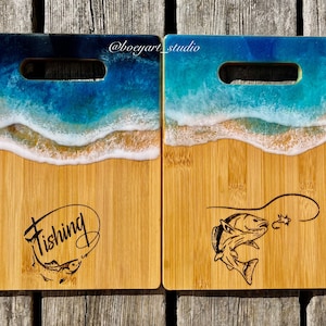 May include: Two wooden chopping boards with a blue and white epoxy resin ocean wave design. One board has the word "Fishing" with a fish and fishing line engraved on it. The other board has a fish jumping out of the water with a fishing line and hook engraved on it.