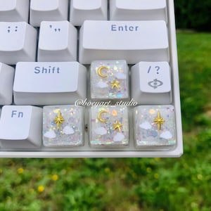 Peut inclure: Gros plan sur un clavier blanc avec des touches personnalisées. Les touches présentent un motif clair et scintillant avec des lunes, des étoiles et des nuages dorés. Les touches Entrée, Maj et autres sont blanches.