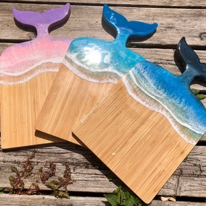 May include: Three wooden serving boards with whale tail handles. The boards feature a beach scene design with resin waves in shades of blue, white, and pink. The whale tails are in shades of purple, blue, and navy.