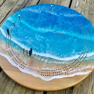 Op de afbeelding: Een rond houten cribbage-bord met een blauw en wit epoxyhars ontwerp van een oceaan golf. Het bord heeft een witte golfkam en een blauwe oceaan achtergrond. Het bord heeft een @boeyart_studio watermerk.