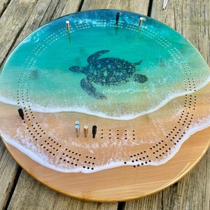 Può includere: Un tabellone da cribbage in legno con una scena di spiaggia e un design a forma di tartaruga marina. Il tabellone è dipinto con acqua blu e verde, sabbia bianca e una tartaruga marina nera. Il tabellone ha un bordo bianco ed è decorato con pioli in metallo dorato.
