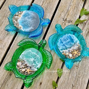 Könnte beinhalten: Drei Schildkrötenförmige Schalen aus Harz mit einer Strandszene im Inneren. Die Schalen sind blau, grün und türkis. Jede Schale hat einen kleinen Seestern, Muscheln und weißen Sand.