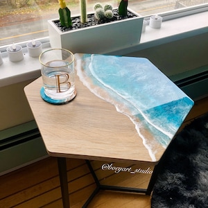 Può includere: Un tavolino esagonale con piano in legno e gambe in metallo nero. Il piano presenta un design a onda oceanica in resina con una spiaggia sabbiosa.