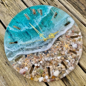 May include: A round resin clock featuring a beach scene. The clock face depicts a turquoise blue ocean with white foamy waves breaking on a sandy beach with seashells and starfish. The clock hands are gold.
