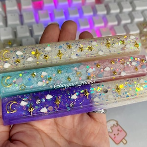 Peut inclure: Trois barres d'espace de clavier en résine colorée avec des motifs célestes. Chaque barre présente des étoiles dorées, des lunes et des paillettes, avec des couleurs allant du blanc au violet. Le texte "@bocygart_studio" est visible.