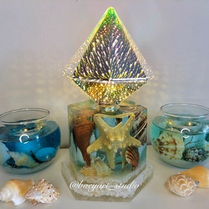 May include: A hexagonal resin lamp base with a starfish and seashells embedded in it. The lamp has a clear diamond-shaped shade with a swirling pattern of light. Two glass bowls with blue water and seashells are on either side of the lamp. The text "@boeyart_studio" is on the base of the lamp.