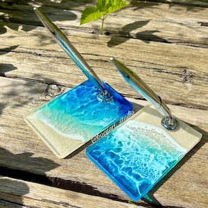 May include: Two pen holders with a beach-themed design. Each holder features a square base with a resin ocean scene in shades of blue and white, topped with a silver pen. The bases have a sandy shore effect. The text "@boogart studio" is visible.