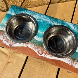 Può includere: Una doppia ciotola per cani con un design a tema spiaggia. La base in legno presenta una scena di spiaggia dipinta con una riva sabbiosa, onde oceaniche e due ciotole in acciaio inossidabile. Le ciotole sono inserite nella base di legno.