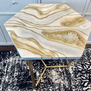 May include: A hexagonal table with a white and gold resin top featuring a swirling, agate-like design with embedded crystals. The table has a gold metal base with a geometric design.
