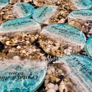 Puede incluir: Primer plano de varios posavasos redondos de resina transparente con un diseño de playa. Cada posavasos presenta un diseño de ola de océano azul y blanco con espuma blanca, arena marrón y guijarros blancos y marrones. El texto "Crystal & Alicia Est. 2022" está impreso en cada posavasos.