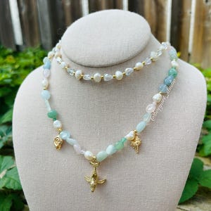 May include: A layered necklace with a delicate gold chain and a longer strand of pastel green, blue, and pink beads. The necklace features a gold charm of a dolphin and two smaller shell charms.