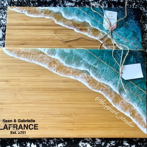 Peut inclure: Deux planches à découper en bois avec un motif d'onde océanique en résine. La planche du haut porte une étiquette indiquant "Fait main avec amour". La planche du bas porte le texte "Sean & Gabrielle LAFRANCE Est. 2021" et "@boogyart_studio" gravé dessus.