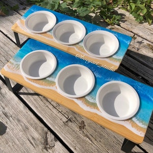Puede incluir: Dos cuencos elevados para mascotas con un diseño de playa. Los cuencos son de cerámica blanca y están colocados sobre una base de madera con un diseño de ola de océano de resina azul y blanca. La base está sostenida por patas de metal negro.