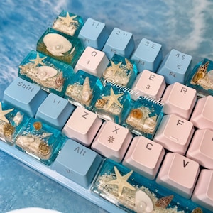 May include: A light blue and pink mechanical keyboard with keycaps featuring ocean-themed resin art. The keycaps have embedded seashells, sand, and starfish. The keyboard has the following keys: Shift, 1!, 2", 3£, 45, €, F4, F1, F2, F3, Q, E, R, T, 3, 11, @botyart studio, F, G, Z, X, C, V, B, Alt.