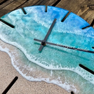 Op de afbeelding: Een ronde wandklok met een strandthema. De wijzerplaat toont een zandstrand, schuimende golven en blauwtinten die de oceaan voorstellen. Zwarte wijzers en de tekst "@boegart_studio" zijn zichtbaar. De klok is gemaakt van hars.