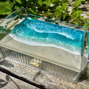 May include: Clear acrylic tray with a beach scene design. The tray features a sandy base with a turquoise and white wave pattern, and gold-colored handles. The tray is rectangular with a ribbed exterior.