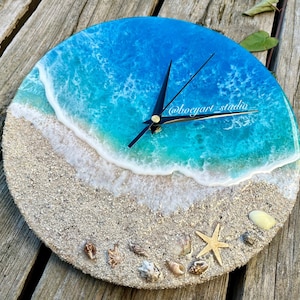 Op de afbeelding: Een ronde klok met een blauw en wit harsontwerp dat een strandscene voorstelt met golven die op de kust breken. De wijzerplaat van de klok is omgeven door wit zand en schelpen. De wijzers van de klok zijn zwart en de wijzerplaat is wit met zwarte cijfers.
