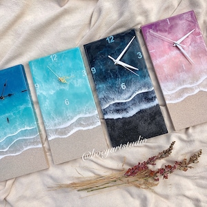May include: Four resin clocks with ocean wave designs. The clocks have white hands and black numbers. The clocks are set on a beige fabric background. The clocks are all different colors: blue, teal, black, and pink.