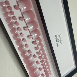 Könnte beinhalten: Ein Set mit 10 pink-weißen Ombre-Press-on-Nägeln in einer durchsichtigen Plastikbox. Die Box hat einen schwarzen Rand und der Text "Pressed Nails By Jen 10/Appliquees" ist auf der Seite aufgedruckt.