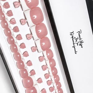 May include: A set of press-on toenails in a French manicure style with a soft pink base and white tips, arranged in a black and white box. The box features the text "Pressed Mijas By Joa" and "19 @pressedmijasbyjoa" printed in black script.