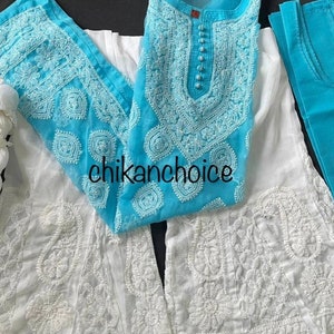 May include: A white and turquoise embroidered dress with a sleeveless top and a long skirt. The dress has a round neckline and a button closure. The skirt is flared and has a scalloped hem. The dress is made of a sheer fabric and is decorated with intricate embroidery.