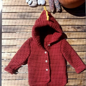 Op de afbeelding: Een rode gehaakte hooded cardigan met een gele dinosaurusvormige capuchon. De cardigan heeft een knoopsluiting en is gemaakt met een textuur.