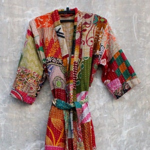 May include: A colorful patchwork robe with a tie belt. The robe features a variety of patterned fabrics in shades of red, orange, green, and pink. The robe is hanging on a wall.