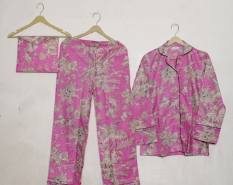 May include: A pink floral pajama set, including a long-sleeved button-down shirt, pants, and a matching pouch, all hanging on wooden hangers. The set features a repeating pattern of beige and blue floral designs with black trim.