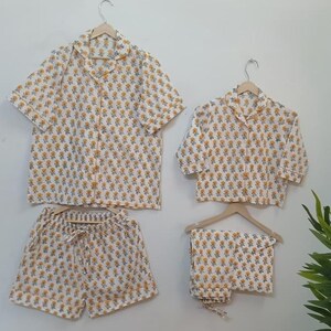 Könnte beinhalten: Vierteiliges Pyjama-Set mit Blumenmuster. Das Set beinhaltet ein kurzärmeliges Hemd, ein kurzärmeliges Oberteil, Shorts und eine lange Hose. Die Pyjamas sind weiß mit einem sich wiederholenden Muster aus gelben Blumen.