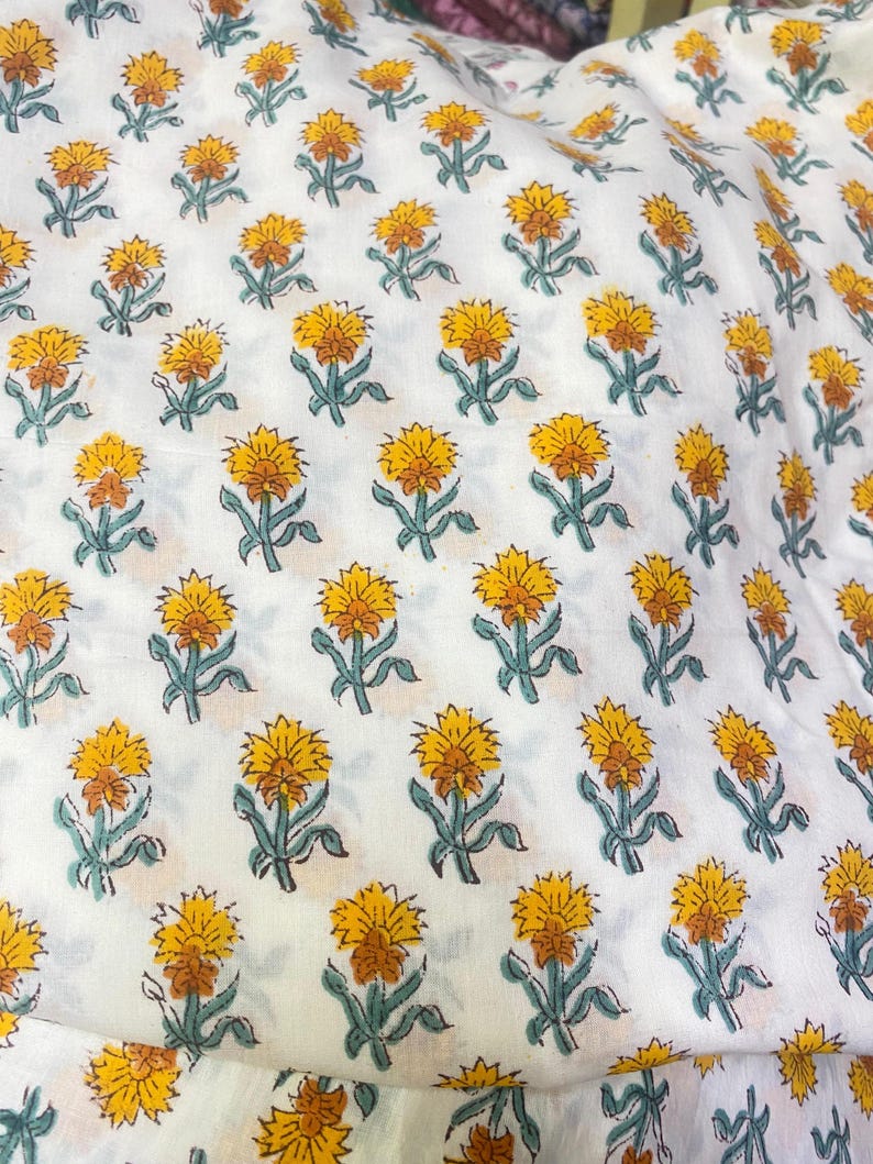 Op de afbeelding: Een witte stof met een herhalend patroon van kleine, gestileerde bloemen met gele bloemblaadjes, oranje harten en gedempte groene stengels en bladeren. De bloemenprint is in een consistent raster over het gehele textieloppervlak geplaatst.