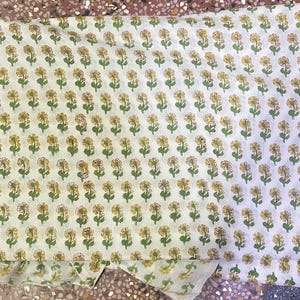 Op de afbeelding: Een gevouwen stuk witte stof met een herhalend patroon van kleine gele bloemen met groene stengels en bladeren. De bloemenprint is gerangschikt in consistente horizontale en verticale rijen over het gehele oppervlak van het textiel.